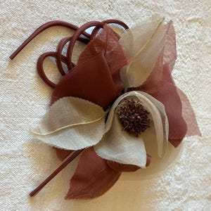 May include: A decorative floral accessory featuring layered fabric petals in shades of terracotta and off-white. The center is composed of a cluster of dark brown stamens, accented by curved, smooth terracotta-colored stems extending outward.