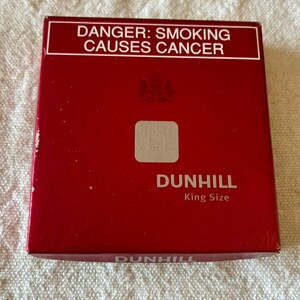 May include: A square, deep red Dunhill King Size cigarette pack featuring a white rectangular box at the top with the text DANGER: SMOKING CAUSES CANCER. The centre displays a small square emblem and the brand name Dunhill in white lettering.