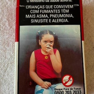Może przedstawiać: Prostokątna paczka papierosów z ostrzeżeniem zdrowotnym o treści O Ministério da Saúde adverte: CRIANÇAS QUE CONVIVEM COM FUMANTES TÊM MAIS ASMA, PNEUMONIA, SINUSITE E ALERGIA. Przedstawia dziecko używające inhalatora oraz ikonę zakazu palenia.