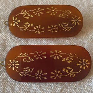 Pode incluir: Duas presilhas de cabelo ovais marrons com desenhos florais dourados. Cada presilha apresenta uma série de flores e folhas douradas, criando um padrão decorativo. As presilhas têm um fecho de metal curvo em uma extremidade.
