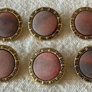 May include: A set of six circular buttons, each featuring a smooth, dusty rose-colored center with a mottled texture. The buttons are framed by a decorative, textured gold-tone metal border with a repeating geometric pattern around the edge.