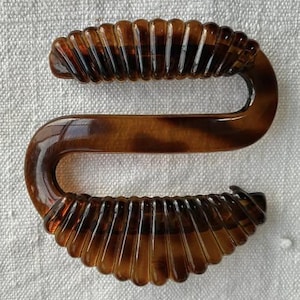 May include: A brown, S-shaped hair clip with a tortoiseshell pattern. The clip has a ribbed design on the top and bottom sections, and a smooth, curved middle section. This hair accessory is suitable for various hairstyles.