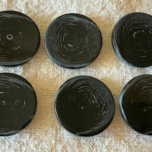 Puede incluir: Un conjunto de seis botones negros circulares, cada uno con un patrón de remolinos blancos que recuerda a la veta de la madera o a anillos concéntricos. Los botones tienen una superficie lisa y pulida, dispuestos en dos filas de tres.