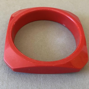 May include: A vibrant red square bangle bracelet. The bracelet has a smooth, glossy finish and a geometric design. The bracelet is approximately 2.5 inches wide and 0.5 inches thick. This bold accessory is perfect for adding a pop of color to any outfit.