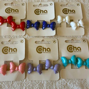 Puede incluir: Seis juegos de dos pinzas para el cabello, cada uno con un diseño de lazo brillante en rojo, azul, blanco, rosa, morado y turquesa. Cada par está montado en una tarjeta blanca de la marca Eha International con un asa recortada superior.