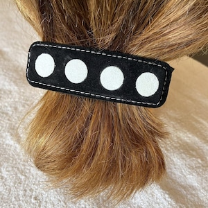Pode incluir: Uma presilha de cabelo preta de camurça com quatro bolinhas brancas e costuras brancas. O acessório de cabelo retangular está segurando uma seção de cabelo castanho. Um acessório de cabelo elegante.