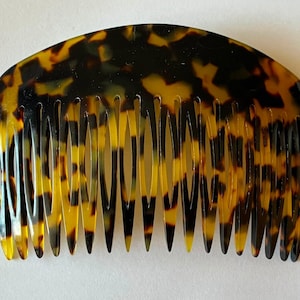 May include: A hair comb featuring a curved top edge and a series of long, tapered teeth. The accessory displays a mottled pattern of dark brown, black, and translucent amber tones, creating a classic tortoiseshell appearance across its surface.