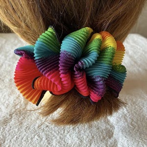 Pode incluir: Uma colorida scrunchie arco-íris feita de tecido plissado. A scrunchie apresenta um espectro de cores, incluindo vermelho, laranja, amarelo, verde, azul e roxo. A scrunchie está enrolada em um rabo de cavalo.