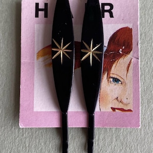 Peut inclure: Deux barrettes à cheveux noires ornées d'étoiles dorées. Elles sont posées sur une carte rose avec le mot "HAIR" et une illustration vintage d'un visage de femme. Accessoire de coiffure rétro.