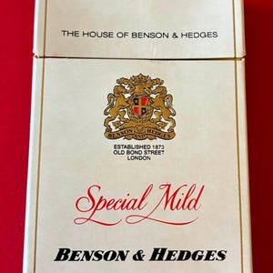 Może przedstawiać: Prostokątna biała paczka papierosów z napisem THE HOUSE OF BENSON & HEDGES u góry. Poniżej znajduje się złoty herb z dwoma lwami, tekst ESTABLISHED 1873 OLD BOND STREET LONDON, czerwony napis Special Mild oraz nazwa BENSON & HEDGES.