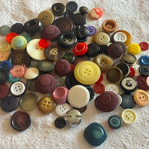 May include: A large collection of assorted sewing buttons in various shapes, sizes, and colors including red, yellow, blue, brown, black, white, and green. The buttons feature different textures, materials, and hole counts, scattered together.