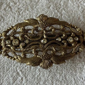 Pode incluir: Broche ovalado em tom dourado antigo com um design floral ornamentado. O broche apresenta um padrão de filigrana com strass redondos e transparentes. O design inclui flores e folhas estilizadas, com um acabamento texturizado e detalhado. O broche tem cerca de 7.6 cm de comprimento.