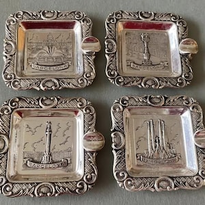 Può includere: Quattro posacenere argentati con cornici quadrate ornate. Ogni vassoio presenta un diverso punto di riferimento di Barcellona, tra cui la Sagrada Familia e una fontana, con il nome della città iscritto sotto. Ogni posacenere ha un piccolo riposo arrotondato.
