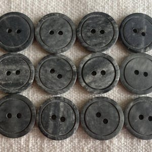 Puede incluir: Doce botones redondos grises con dos agujeros cada uno, dispuestos en tres filas de cuatro. Los botones tienen un aspecto veteado y un anillo exterior rebajado. Los botones están sobre una superficie blanca texturizada.