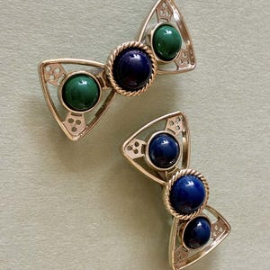 Pode incluir: Duas presilhas de cabelo prateadas em forma de laço. Cada presilha apresenta um cabochão azul escuro central, emoldurado por um design de corda torcida, e ladeado por cabochões verdes e azuis emoldurados em armações triangulares. As presilhas são exibidas em uma superfície verde clara.