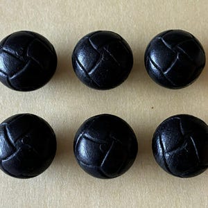 May include: Six black, round buttons with a textured, woven-like pattern. The buttons are evenly spaced and arranged in two rows of three on a light tan surface. The buttons appear to be made of leather or a similar material.