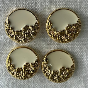 Puede incluir: Cuatro botones redondos de color crema con adornos florales dorados. Los botones tienen un borde dorado y un centro de color crema. El diseño floral está agrupado en la parte inferior de cada botón.