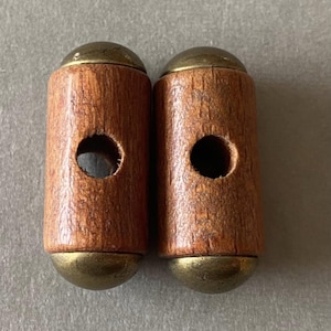 May include: Two wooden toggle clasps with brass-colored metal caps and bases. Each clasp has a central hole. The wood has a warm brown tone, and the metal has a slightly aged appearance.