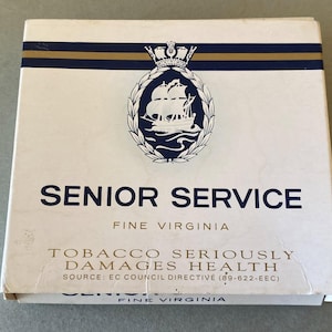 Pode incluir: Uma caixa de cigarros Senior Service branca e azul. A caixa apresenta uma faixa azul marinho e dourada, com a inscrição "SENIOR SERVICE" em azul marinho. Também inclui um emblema de navio e um aviso de saúde.