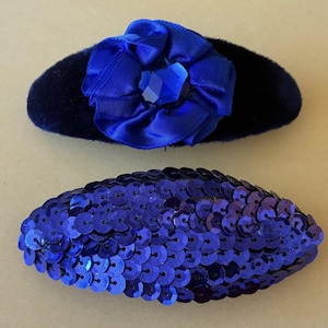 Pode incluir: Duas presilhas de cabelo azuis. A presilha superior é oval, coberta de veludo azul escuro e adornada com uma flor de tecido azul e uma joia central. A presilha inferior é oval e coberta de lantejoulas azuis.
