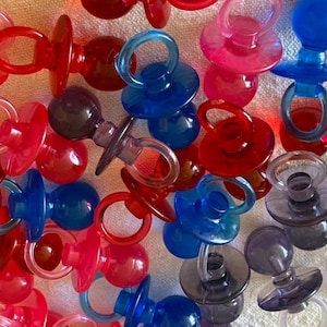 Puede incluir: Una colección de chupetes de plástico translúcido en varios colores, incluyendo rojo, azul, rosa y morado. Cada chupete presenta una tetina redonda, un escudo circular y un pequeño anillo, dispuestos en un conjunto denso y superpuesto.