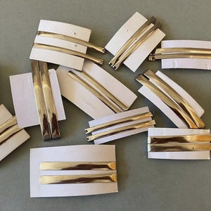 Pode incluir: Uma variedade de grampos e presilhas de cabelo de metal dourado, exibidos em embalagens de cartão branco. Os grampos variam em estilo, incluindo designs curvos e retos. O fundo é cinza suave.