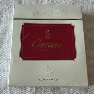 Könnte beinhalten: Eine weiße Zigarettenschachtel mit einem roten rechteckigen Etikett mit dem goldenen Text "Cartier PARIS LONDON NEW YORK". Die Schachtel trägt auch die Worte "LUXURY MILD" und dekorative Embleme.