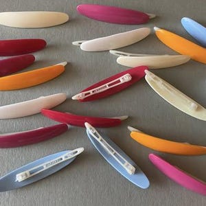 Op de afbeelding: Een verzameling vintage haarspeldjes in verschillende kleuren, waaronder rood, roze, oranje, wit en blauw. De haarspeldjes zijn gemaakt van plastic en hebben een gebogen, langwerpige vorm met een metalen sluiting.