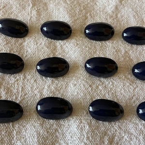 Puede incluir: Doce cabujones ovalados de color azul marino oscuro sobre una tela blanquecina texturizada. Las piedras pulidas están dispuestas en tres filas de cuatro. Cada cabujón tiene una superficie lisa y brillante.