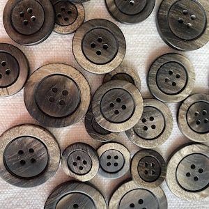 Puede incluir: Una colección de botones grises oscuros de varios tamaños, cada uno con cuatro agujeros. Los botones tienen un aspecto de veta de madera y están dispuestos sobre una superficie blanca. Los botones son redondos con un borde ligeramente elevado.