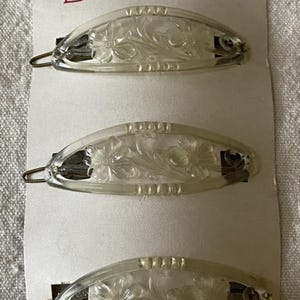 Puede incluir: Tres horquillas ovaladas transparentes con diseños florales, sujetas a una tarjeta blanca. Las horquillas tienen un cierre metálico y están etiquetadas como "color Delcia". La tarjeta también tiene el texto "Modelo y Marca registrados".