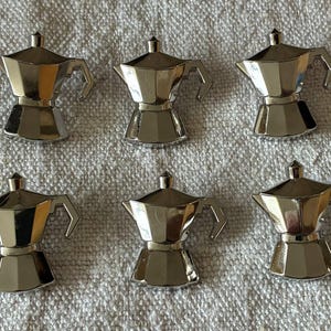 Op de afbeelding: Zes kleine, glanzende zilveren espressomakers op een getextureerd, gebroken wit oppervlak. Elke miniatuur koffiepot heeft een klassiek ontwerp met een handvat en een tuit.