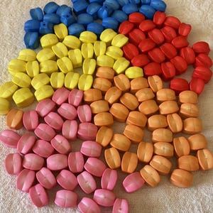 May include: A collection of small, smooth, rounded beads in various colors including blue, yellow, red, orange, and pink. Each bead features a central horizontal indentation and a small hole, arranged in a loose, scattered pile on a textured surface.