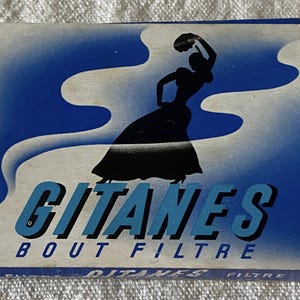 Puede incluir: Una caja de cigarrillos vintage con un diseño azul y blanco. La caja presenta el texto "GITANES BOUT FILTRE" en azul, con una silueta de una mujer con un vestido. La caja es rectangular y tiene un aspecto desgastado.