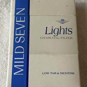 Pode incluir: Um maço de cigarros Mild Seven Lights. A embalagem é predominantemente branca com uma faixa vertical azul no lado esquerdo. O texto "MILD SEVEN" é impresso verticalmente em branco na faixa azul. O texto "Lights" e "CHARCOAL FILTER" estão impressos na frente.