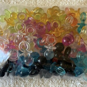 Puede incluir: Una bolsa de plástico transparente llena de una gran colección de pequeños dijes translúcidos con forma de chupete en una variedad de colores que incluyen amarillo, naranja, rosa, morado, azul y negro, cada uno con un anillo y una tetina redondeada.