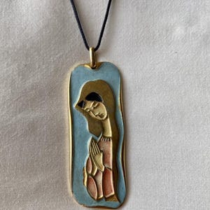 Pode incluir: Um colar com pingente retangular com uma ilustração estilizada de uma figura em oração. O pingente tem um fundo azul claro, uma moldura dourada e um cordão preto.