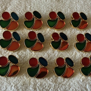 Op de afbeelding: Een verzameling van twaalf kleurrijke, abstract gevormde oorbellen. Elke oorbel heeft een goudkleurige rand met rode, groene, oranje en blauwe emaille secties. De oorbellen liggen op een getextureerd, gebroken witte ondergrond.