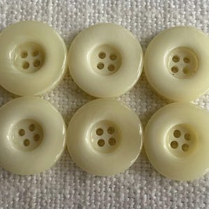 Peut inclure: Six boutons ronds de couleur crème avec une finition légèrement brillante. Chaque bouton a un centre en retrait avec quatre petits trous pour la couture. Les boutons sont disposés en deux rangées de trois sur une surface blanche texturée.