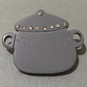 Pode incluir: Broche cinzento em forma de panela com tampa. O broche tem duas pegas e é decorado com strass transparentes ao longo da borda da tampa. O design da panela inclui linhas onduladas.