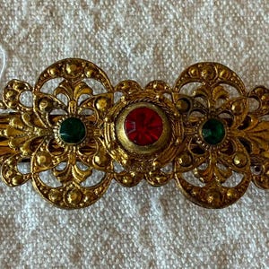 Pode incluir: Uma presilha de cabelo em metal dourado com um design de filigrana intrincado, uma pedra preciosa vermelha circular central ladeada por duas pedras verdes circulares menores. O metal tem um acabamento texturizado com padrões ornamentados.