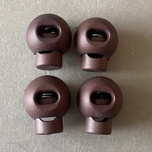 May include: Four brown cord locks with a spherical top and a cylindrical base. Each cord lock has a small opening and a metal spring inside. The cord locks are made of plastic.