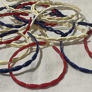 Pode incluir: Uma coleção de tiaras coloridas em vermelho, branco e azul. As tiaras têm um design torcido e são feitas de um material brilhante, semelhante ao plástico. As tiaras estão espalhadas sobre uma superfície branca.