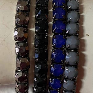 Puede incluir: Cuatro pulseras de cuentas con piedras cuadradas en tonos gris oscuro, negro, azul y gris claro. Las pulseras se muestran una al lado de la otra, mostrando la variedad de colores y texturas.