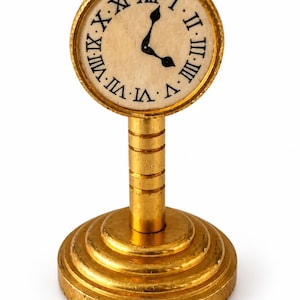 May include: A gold-toned table clock featuring a circular face with black Roman numerals and two black hands. The clock is mounted on a cylindrical pedestal with horizontal grooves, resting on a tiered, circular base with a textured edge.
