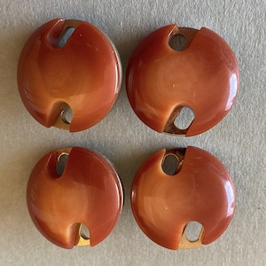 Pode incluir: Quatro botões redondos castanho-avermelhados com um design único. Cada botão tem uma forma circular com um recorte e um verso de metal dourado. Os botões estão dispostos num padrão quadrado.
