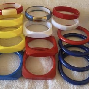 Pode incluir: Uma coleção de pulseiras coloridas em vários designs e cores. O sortido inclui pulseiras redondas, quadradas e estampadas em tons de vermelho, azul, amarelo, branco e verde. Algumas têm listras ou padrões geométricos.