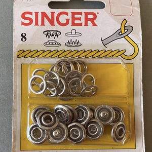 Pode incluir: Um pacote de fechos de pressão da marca Singer. A embalagem branca e amarela apresenta o logotipo Singer e o número 8. Contém fechos de pressão de metal prateado, perfeitos para costura.