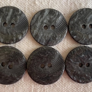 Puede incluir: Seis botones redondos de color gris oscuro con dos agujeros cada uno. Los botones tienen un aspecto veteado y una serie de líneas curvas en la superficie. Los botones miden aproximadamente 3,2 cm de diámetro.