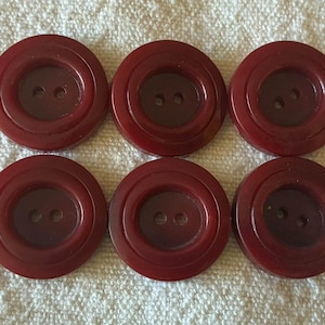 May include: A set of six round, dark red buttons arranged in two rows of three. Each button features a smooth, glossy finish with a recessed center and two small holes for sewing, showcasing a consistent deep burgundy color throughout the set.